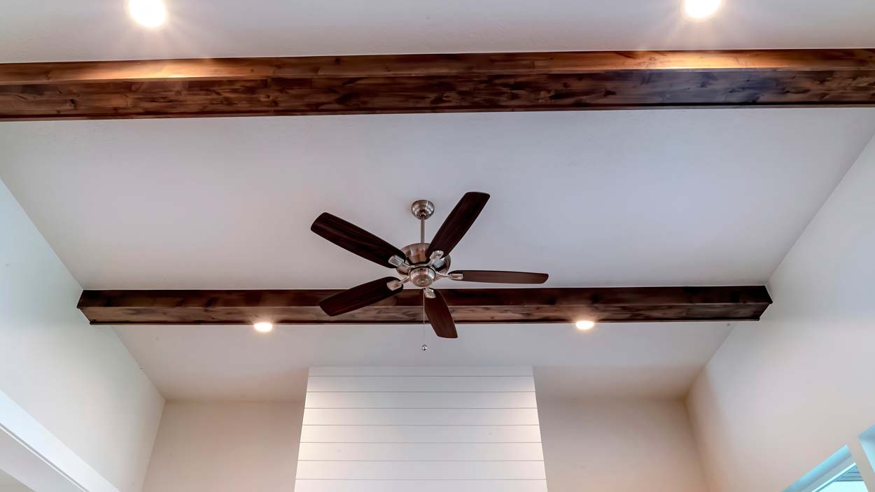 Panorama crop Ceiling fan with lights between decorative wood beams inside living room of home. Recessed round lights can also be seen over the room with white wall.